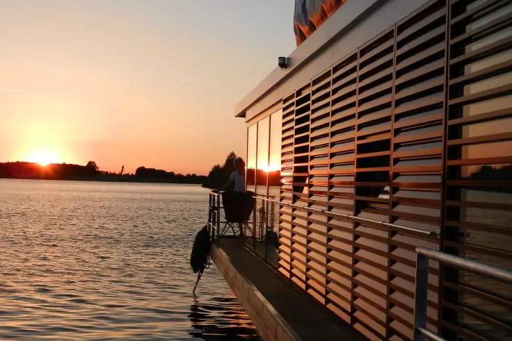 Geb&auml;ude mit horizontalen Lamellen am Ufer, Sonnenuntergang &uuml;ber dem Wasser, zwei Personen auf einem schmalen Steg
