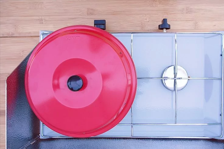 Runder roter Topfdeckel auf der linken Herdplatte eines Gasherds, rechte Herdplatte leer, Holzarbeitsplatte im Hintergrund