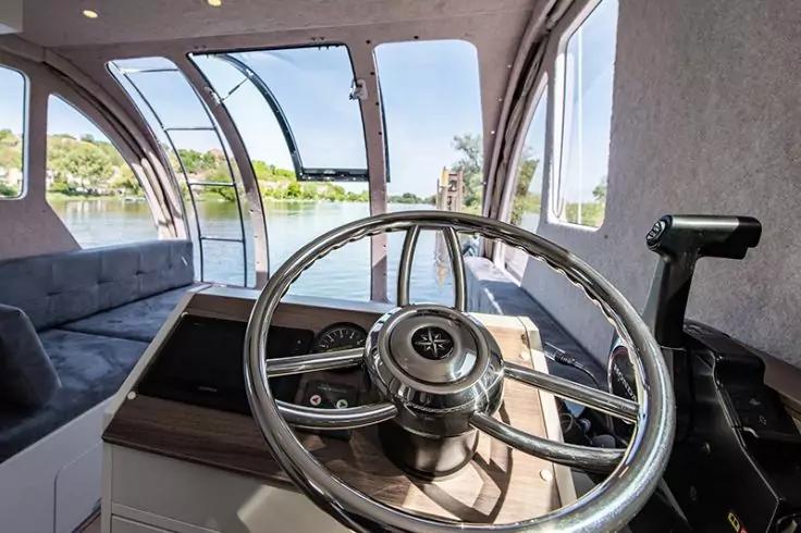 Blick auf das Steuerrad und die Armaturen in einer Bootssteuerkabine mit gebogenen Fenstern, au&szlig;en Wasser, Uferb&auml;ume und Geb&auml;ude