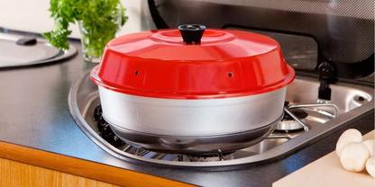 Runder silberner Topf mit rotem Deckel und schwarzem Griff auf einem Gasherd; zwei Knoblauchzehen auf der Arbeitsplatte; gr&uuml;ne Topfpflanze im Hintergrund