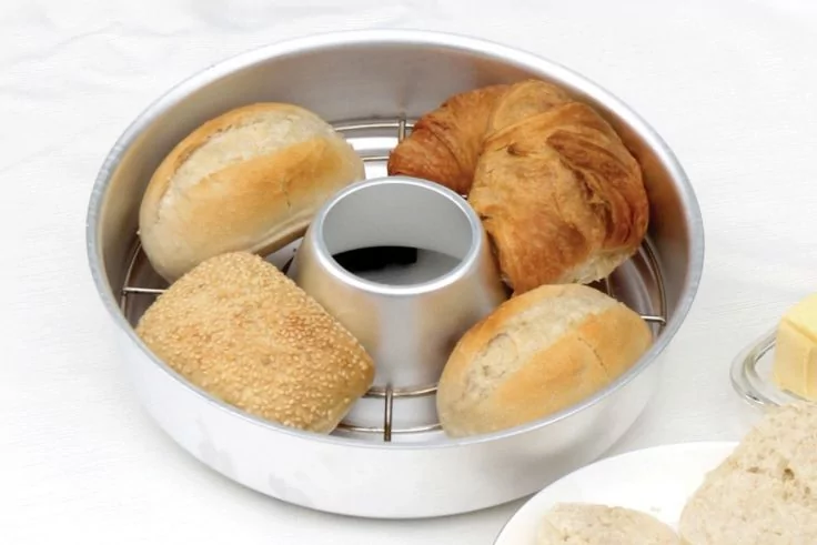 Runde Metallbackform mit Rohr in der Mitte und Rost, darauf drei Br&ouml;tchen, ein Sesambr&ouml;tchen und ein Croissant; rechts Teller mit Butter und aufgeschnittenem Brot