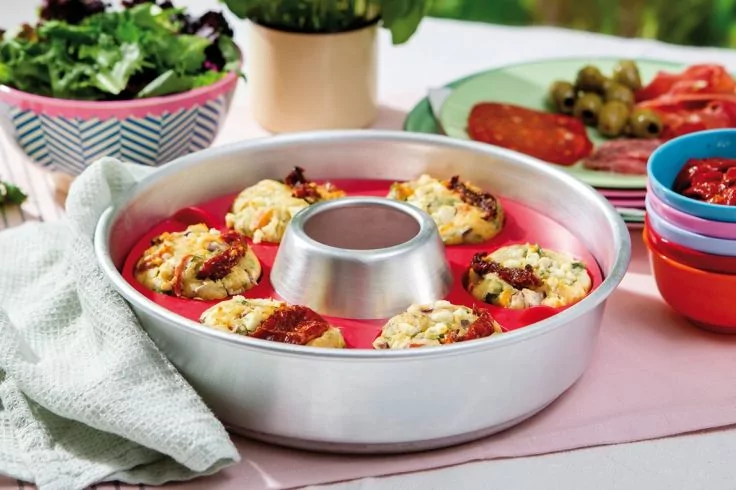 Runde Backform mit Loch in der Mitte, darin sechs gebackene Teigportionen mit K&auml;se und Tomaten; im Hintergrund Salatsch&uuml;ssel, Teller mit Oliven und Tomatenscheiben, Stapel bunter Schalen und eine Stoffserviette