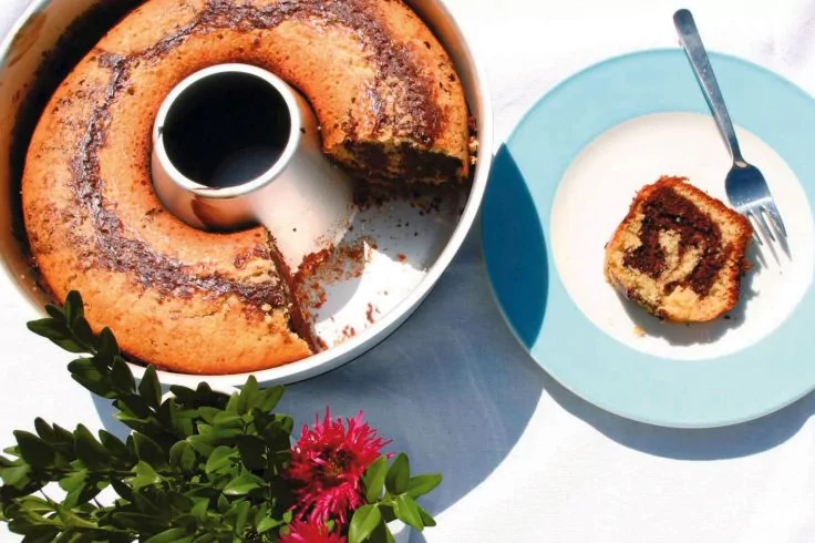 Gugelhupf mit Marmormuster in Backform, ein St&uuml;ck auf blauem Teller, Gabel, gr&uuml;ne Pflanze mit rosa Bl&uuml;ten auf wei&szlig;er Tischdecke