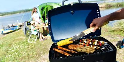 Offener Grill mit Spie&szlig;en, Steak, W&uuml;rstchen und Maiskolben; eine Hand mit Grillzange h&auml;lt den Mais; im Hintergrund Ufer, Wasser und Personen beim Picknick