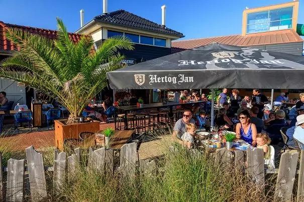 Au&szlig;enbereich eines Caf&eacute;s mit schwarzem Sonnenschirm mit Aufschrift 'Hertog Jan', mehreren Personen an Tischen, Kind, Blumenk&uuml;bel mit Palme, Holzzaun und Gras im Vordergrund, Geb&auml;ude und blauer Himmel im Hintergrund