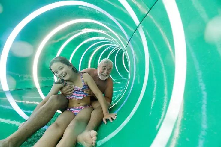 Ein Erwachsener sitzt hinter einem Kind in einer gr&uuml;nen, spiralf&ouml;rmigen Wasserrutsche