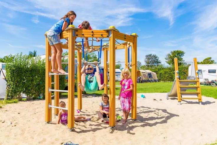 Mehrere Kinder auf einem Holz-Kletterger&uuml;st auf sandigem Untergrund, Wohnwagen und blauer Himmel im Hintergrund