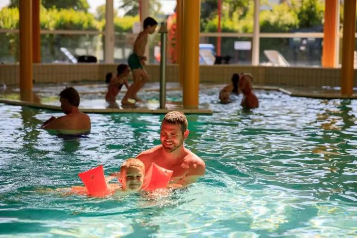 Mann h&auml;lt ein Kind mit orangefarbenen Schwimmfl&uuml;geln im Wasser eines Hallenbads; mehrere Personen und S&auml;ulen mit gro&szlig;en Fenstern im Hintergrund
