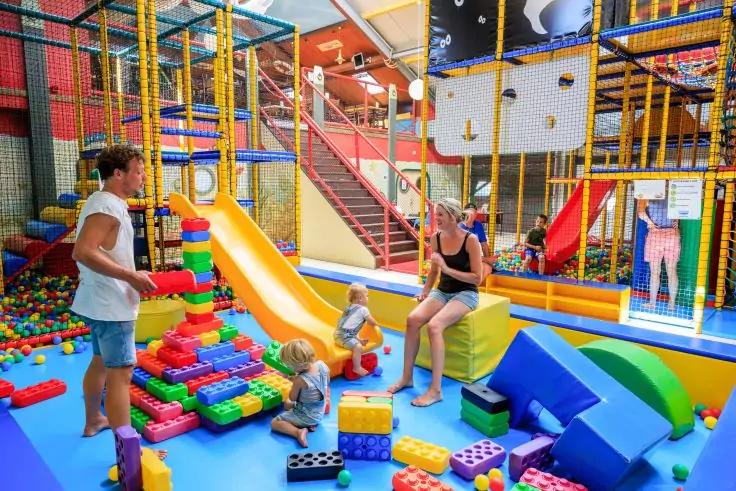 Indoor-Spielplatz mit bunten Schaumstoffbl&ouml;cken, gro&szlig;en Plastikbausteinen, gelber Rutsche und B&auml;llebereich; Kletterger&uuml;st und Netze im Hintergrund; ein Mann steht links, eine Frau sitzt auf einem Block, zwei Kinder spielen auf dem Boden