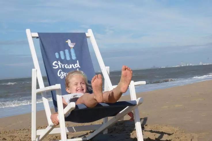 Ein Kind sitzt in einem blauen Strandliegestuhl auf dem Sand, die F&uuml;&szlig;e nach vorn ausgestreckt; im Hintergrund Meer und Himmel; auf dem Stoff sind eine M&ouml;we und der Schriftzug 'Strand gut'