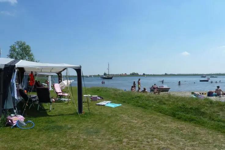 Zelt- oder Sonnenschutz links mit St&uuml;hlen und Gegenst&auml;nden, grasbewachsene Fl&auml;che mit Handt&uuml;chern, mehrere Personen am Ufer und im Wasser, Boote auf dem Wasser, weiter Horizont unter blauem Himmel