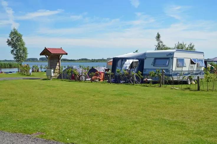 Wohnwagen mit ausgefahrener Markise und mehreren Fahrr&auml;dern auf einer Rasenfl&auml;che, daneben ein kleiner h&ouml;lzerner Unterstand; im Hintergrund ein See, B&auml;ume und blauer Himmel