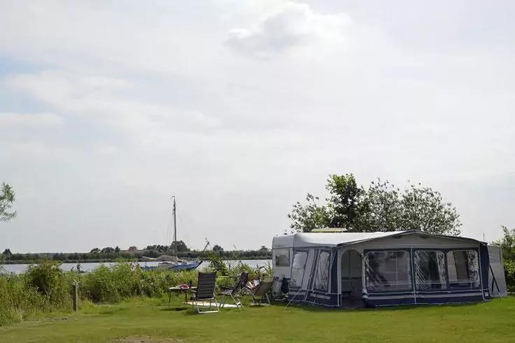 Wohnwagen mit Vorzelt auf einer Wiese am See, zwei Klappst&uuml;hle und ein kleiner Tisch, B&auml;ume und Boote im Hintergrund, bew&ouml;lkter Himmel