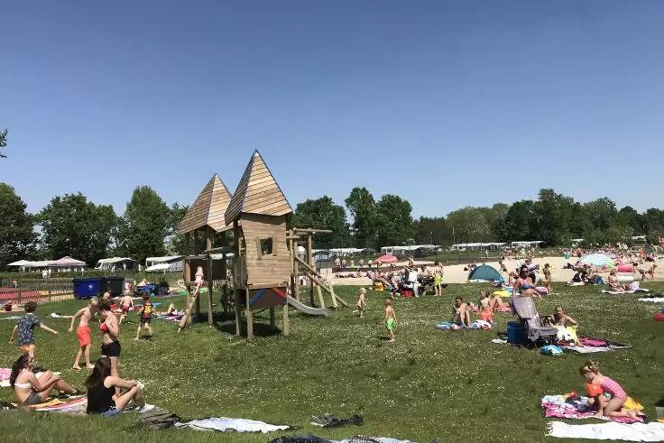 Holz-Spielger&uuml;st mit zwei spitzen D&auml;chern auf einer Grasfl&auml;che, umgeben von Menschen auf Handt&uuml;chern und unter Sonnenschirmen, Wasser und B&auml;ume im Hintergrund, blauer Himmel