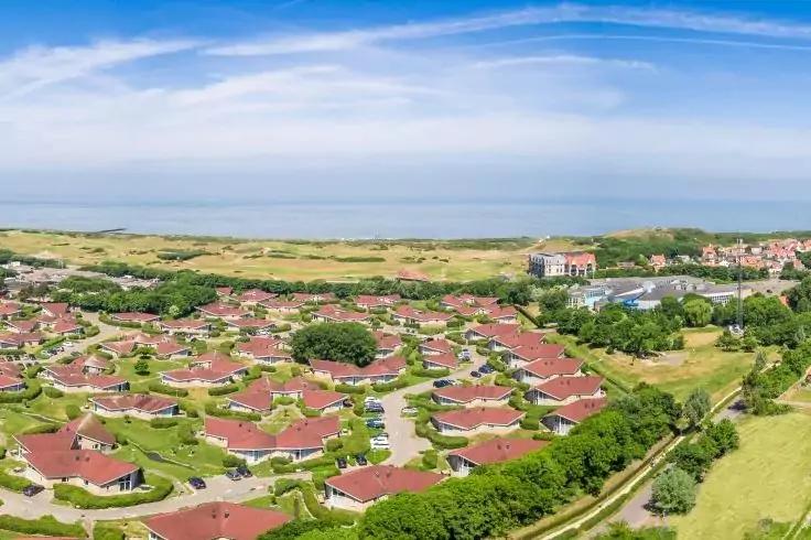 Luftaufnahme zahlreicher einzelner H&auml;user mit roten D&auml;chern in kreisf&ouml;rmigen Anordnungen, mit Gr&uuml;nfl&auml;chen, Stra&szlig;en und geparkten Autos; Meer am Horizont und blauer Himmel mit Wolken
