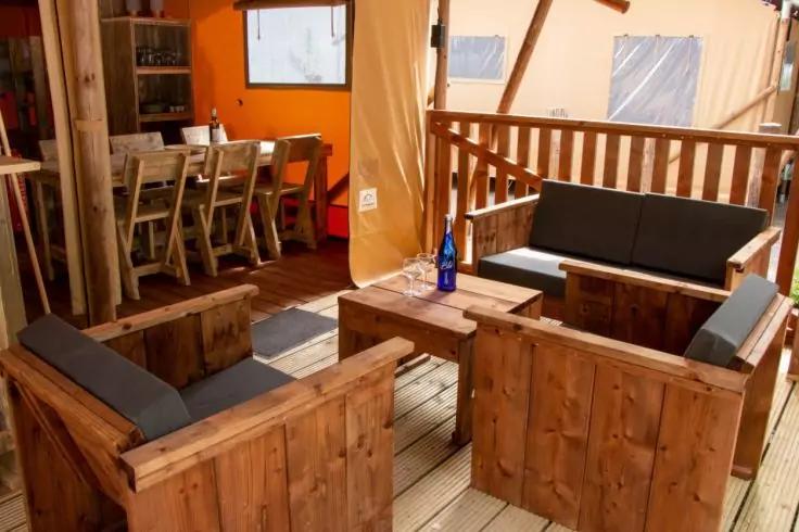 &Uuml;berdachte Terrasse mit Holzboden, zwei Holzsessel, eine Holzbank mit dunkelgrauen Kissen, niedriger Holztisch mit blauer Flasche, Holzgeländer und Esstisch mit St&uuml;hlen im Hintergrund