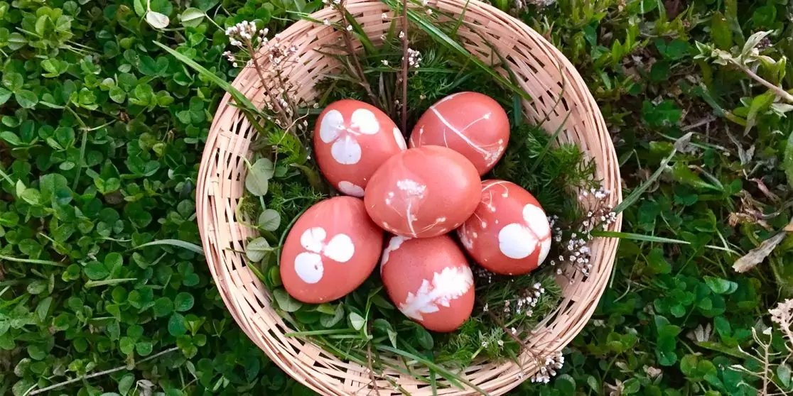 Runder geflochtener Korb auf gr&uuml;nem Gras mit sechs rot-rosa Eiern mit wei&szlig;en Blumenmustern, kleinen Zweigen und kleinen wei&szlig;en glockenf&ouml;rmigen Bl&uuml;ten