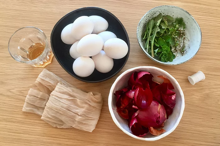 Sechs wei&szlig;e Eier in einer schwarzen Schale, rote Zwiebelschalen in einer wei&szlig;en Schale, kleine Schale mit gr&uuml;nen Kr&auml;utern, Glas mit bernsteinfarbener Fl&uuml;ssigkeit, gefaltetes hellbeiges Tuch und kleiner wei&szlig;er Zylinder auf einer Holzoberfl&auml;che