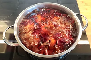 Edelstahltopf auf Cerankochfeld mit braunroter, kochender Fl&uuml;ssigkeit und schwimmenden roten Zwiebelschalen; rechts ein Holzbrett