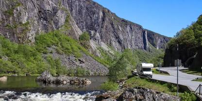 Fluss mit Felsen und flie&szlig;endem Wasser, rechts ein parkendes Wohnmobil an einer Stra&szlig;e, links eine steile Felswand und bewaldete H&auml;nge, blauer Himmel