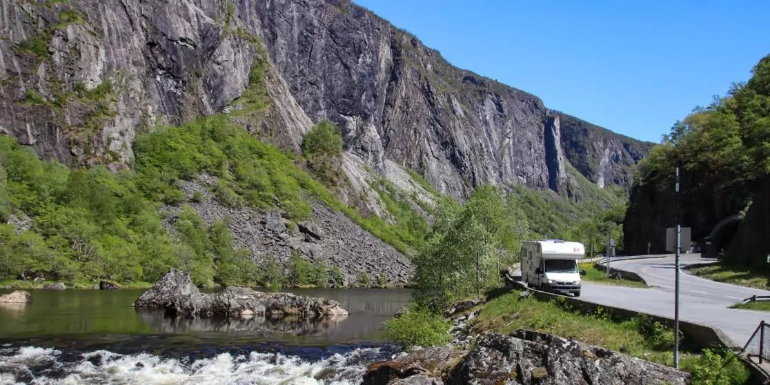 Fluss mit Felsen und flie&szlig;endem Wasser, rechts ein parkendes Wohnmobil an einer Stra&szlig;e, links eine steile Felswand und bewaldete H&auml;nge, blauer Himmel