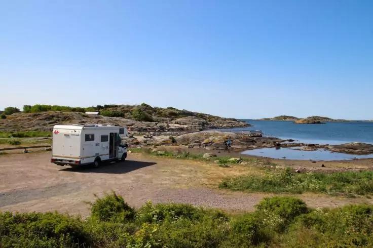 Wei&szlig;es Wohnmobil auf Schotterplatz neben felsiger K&uuml;ste und ruhigem Meer, blauer Himmel, niedrige Vegetation im Vordergrund
