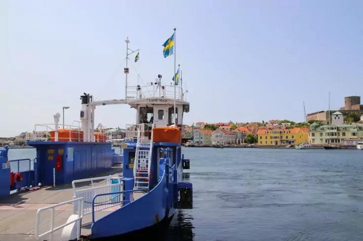 Blau-wei&szlig;e F&auml;hre mit abgesenkter Rampe am Kai, Wasser im Vordergrund, bunte H&auml;user und ein Turm am Ufer, schwedische Flagge am Mast