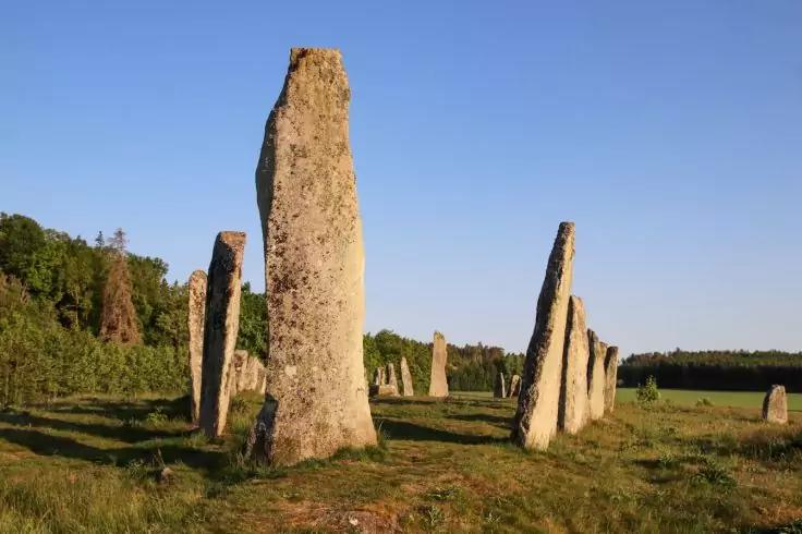 Mehrere aufrecht stehende Steine auf einer Wiese, B&auml;ume im Hintergrund, blauer Himmel