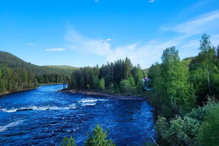 Fluss mit kleinen Stromschnellen, bewaldete Ufer mit Nadel- und Laubb&auml;umen, blauer Himmel mit vereinzelten Wolken, kleines Haus am rechten Ufer