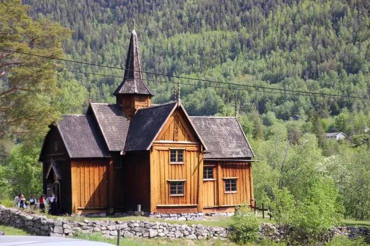 Holzgeb&auml;ude mit spitzem Turm und mehreren Satteld&auml;chern, Steinmauer im Vordergrund, kleine Gruppe Menschen links, Wiese und B&auml;ume, bewaldeter Hang im Hintergrund, Stromleitungen quer durchs Bild