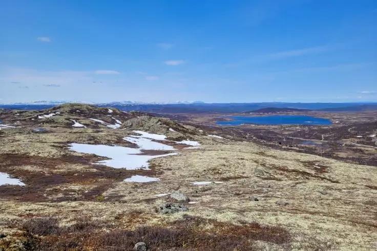 H&uuml;gellandschaft mit sp&auml;rlicher Vegetation, Steinen, Schneefeldern, einem See in der Ferne und blauem Himmel mit wenigen Wolken