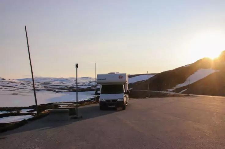 Wei&szlig;es Wohnmobil auf asphaltierter Parkfl&auml;che vor schneebedeckten Bergen, tief stehende Sonne am rechten Bildrand, mehrere Masten sichtbar