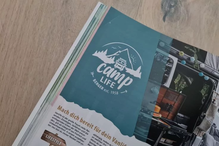 Aufgeschlagenes Magazin auf Holztisch, linke Seite blau mit wei&szlig;em 'Camp Life'-Logo und Bergmotiv, rechte Seite Foto einer Wohnmobil-Innenansicht und Text