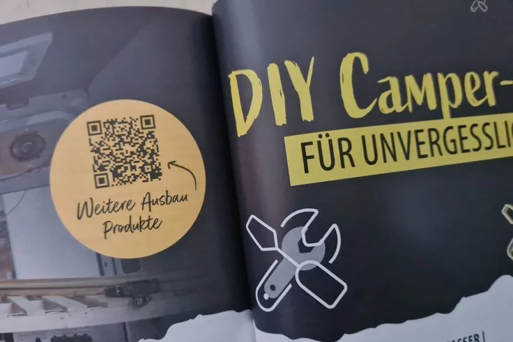 Aufgeschlagene Magazinseite mit dem Text 'DIY Camper' und der teilweisen Zeile 'F&Uuml;R UNVERGESSL...'; Schraubenschl&uuml;ssel-Symbole unten rechts; runder Aufkleber links mit QR-Code und handschriftlichem 'Weitere Ausbau Produkte'