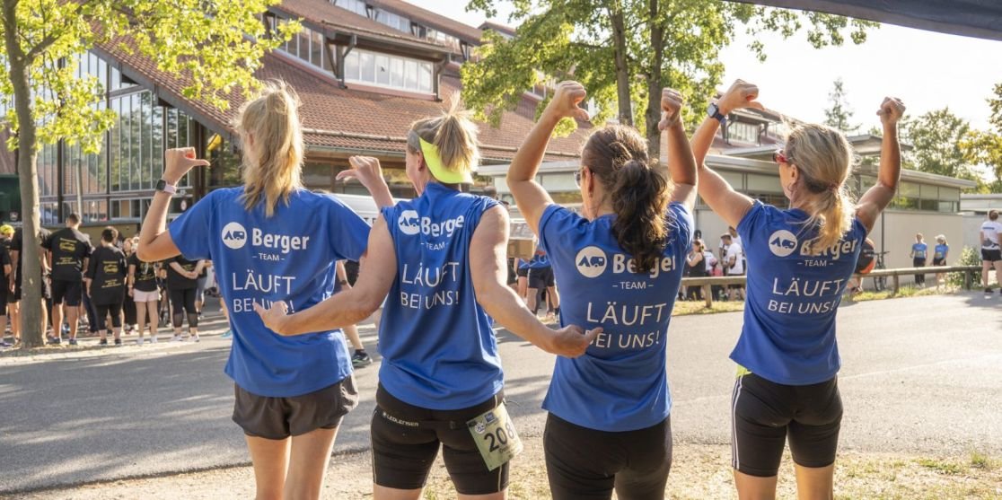 Vier Personen von hinten, blaue T-Shirts mit Aufdruck 'Berger' und 'L&Auml;UFT BEI UNS', schwarze Shorts, erhobene Arme, Stra&szlig;e mit B&auml;umen und Geb&auml;uden im Hintergrund
