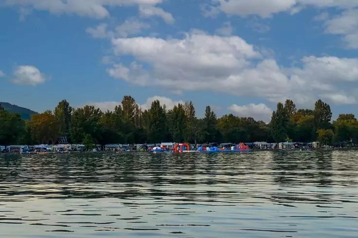 Wasser mit Spiegelungen im Vordergrund; am Ufer parkende Autos und bunte Zelte; Baumreihe und Himmel mit vereinzelten Wolken