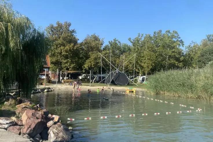 See mit einer Linie aus Bojen, mehrere Personen stehen im flachen Wasser, Felsen am linken Ufer, Schilf rechts, B&auml;ume und ein kleines Geb&auml;ude im Hintergrund, blauer Himmel