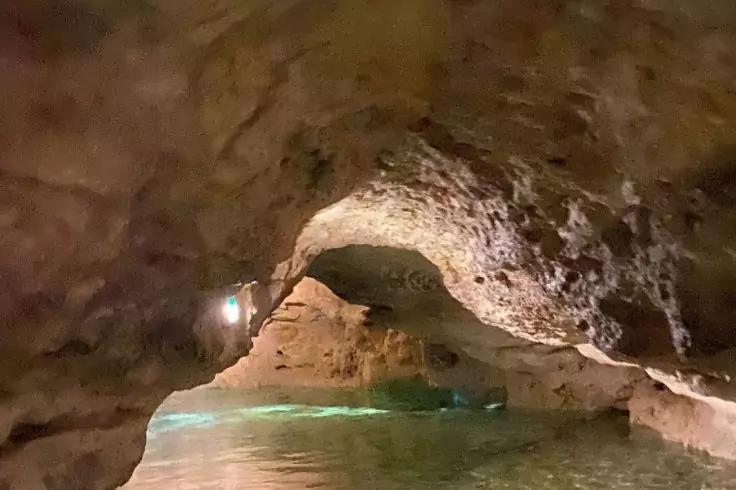 Inneres einer Felsenh&ouml;hle mit niedrigem gew&ouml;lbtem Deckenbogen &uuml;ber seichtem Wasser, gr&uuml;nliche Spiegelung auf der Wasseroberfl&auml;che und eine kleine Lichtquelle an der linken Wand