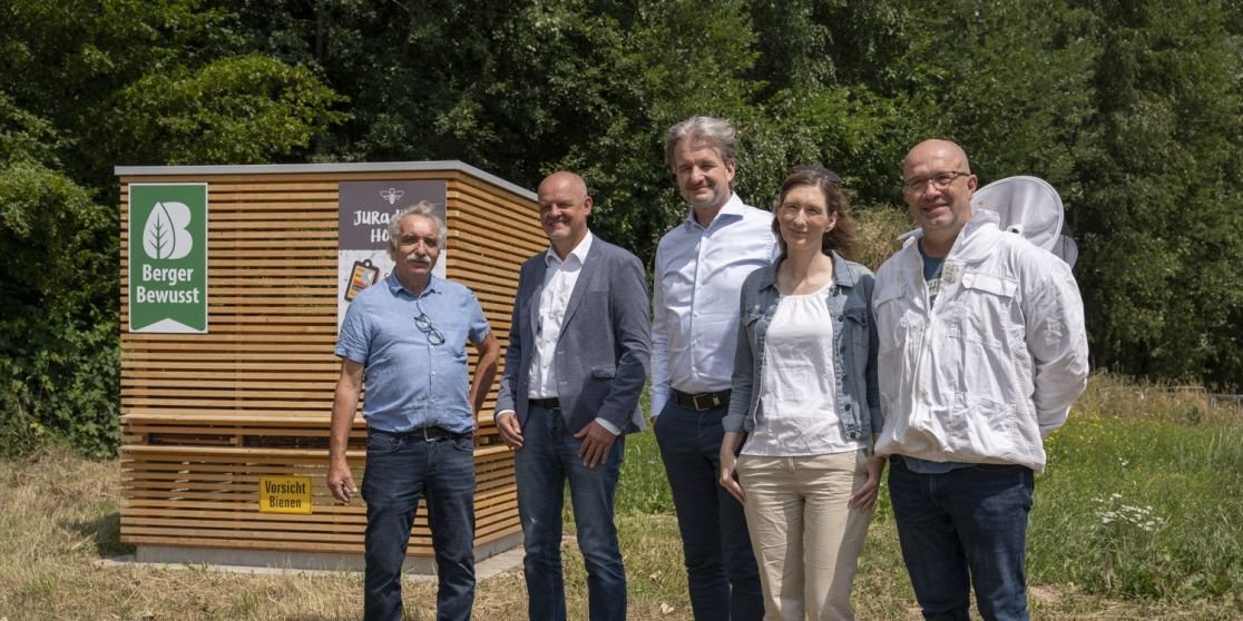F&uuml;nf Personen stehen nebeneinander vor einem h&ouml;lzernen Informationskasten mit Schildern auf einer Wiese, dahinter B&auml;ume