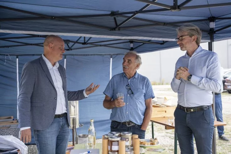 Drei M&auml;nner unter einem blauen Pavillon; links ein Mann in grauem Sakko gestikulierend, Mitte ein &auml;lterer Mann in hellblauem Kurzarmhemd mit einem Glas in der Hand, rechts ein Mann mit Brille und wei&szlig;em Hemd mit zusammengehaltenen H&auml;nden; vor ihnen e