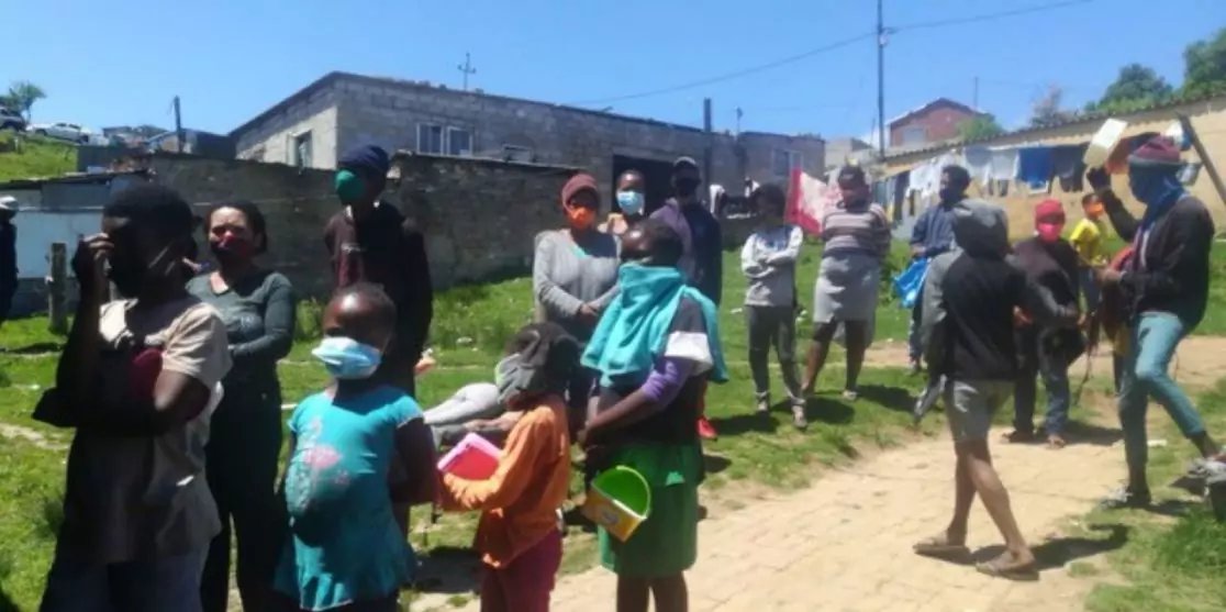 Mehrere Personen, darunter Kinder und Erwachsene, stehen im Freien auf einem sandigen Weg neben Gras und Geb&auml;uden; einige tragen Mund-Nasen-Schutz und halten Plastikschalen oder Eimer