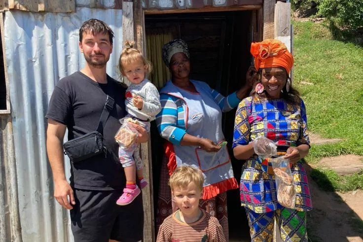 Fünf Personen vor einer Wellblechhütte mit offenem Holztürrahmen: ein Mann in schwarzem T‑Shirt mit Umhängetasche hält ein Kleinkind mit pinken Schuhen; ein Junge steht vorne; zwei Frauen in bunter Kleidung und Kopfbedeckungen, eine mit Schürze hält