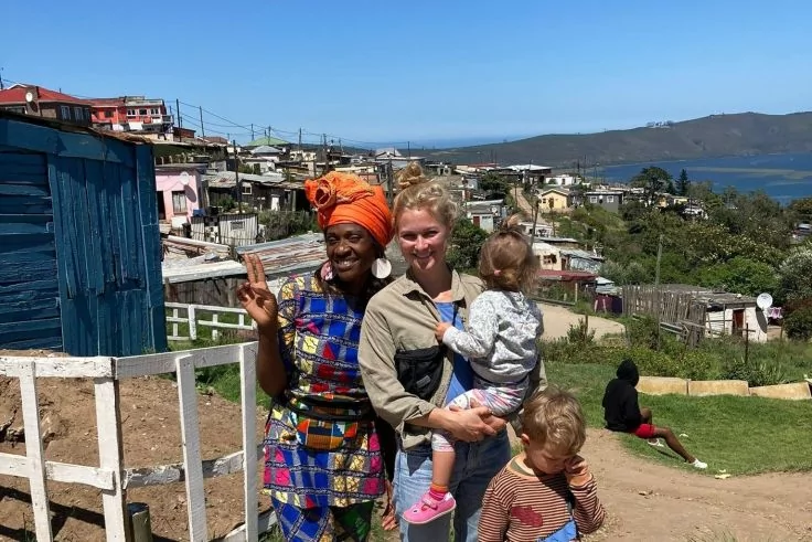 Zwei Frauen stehen vor Häusern auf einem Hügel mit Meer im Hintergrund. Eine trägt ein buntes Kleid und ein oranges Kopftuch und macht ein Peace-Zeichen; die andere trägt ein Kleinkind in einer Trage. Ein weiteres Kind steht davor
