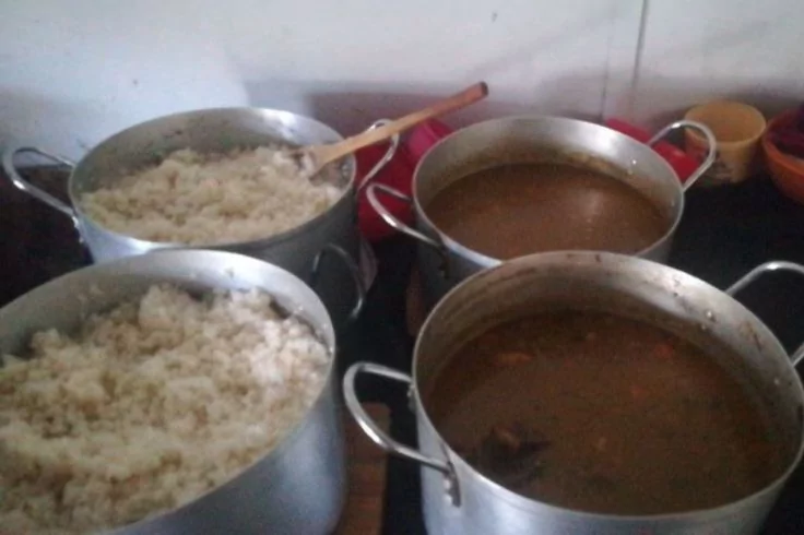 Vier große Metalltöpfe auf einer Arbeitsfläche; zwei mit weißem Reis, zwei mit brauner Flüssigkeit. Ein Holzlöffel in einem Reis-Topf; mehrere Tassen im Hintergrund