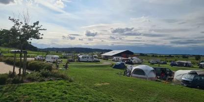 Campingplatz mit mehreren Zelten, Wohnwagen und Autos auf einer Wiese, flaches Geb&auml;ude, B&auml;ume, H&uuml;gel in der Ferne und bew&ouml;lkter Himmel