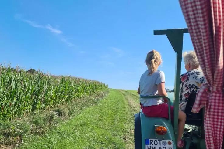 Zwei Personen sitzen auf einem kleinen blauen Traktor auf einem grasigen Weg zwischen einem Maisfeld und einer Wiese; rote kariere Vorhangkante rechts, blauer Himmel