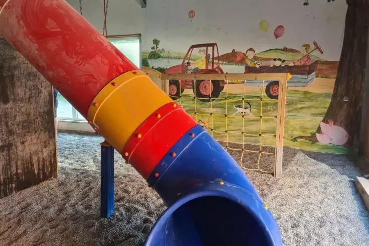 Bunte R&ouml;hrenrutsche (rot, gelb, blau) in einer Innen-Spielzone &uuml;ber losem Bodenmaterial, Kletternetz, Holzpfosten und Wandbild mit Traktor und Tieren