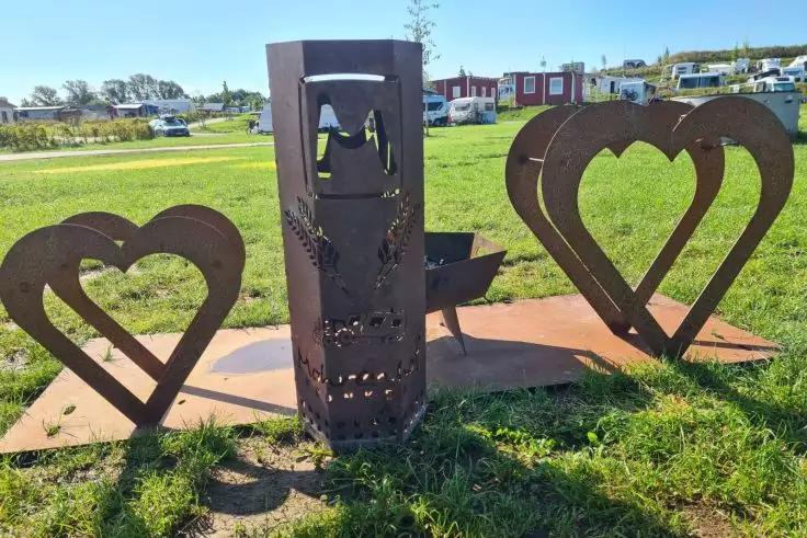 Drei rostbraune Metallskulpturen auf einer Metallplatte im Gras: links und rechts je ein herzf&ouml;rmiger Rahmen, mittig ein hohes rechteckiges Objekt mit Ausstanzungen; Wohnwagen und Geb&auml;ude im Hintergrund, blauer Himmel