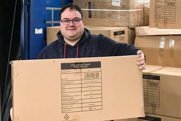 Person h&auml;lt gro&szlig;en Versandkarton vor Lager mit gestapelten Kartons