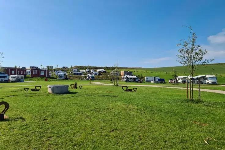 Gr&uuml;ne Wiese mit mehreren Wohnmobilen und Wohnwagen in einer Reihe, kleinen B&auml;umen, runder Betonstruktur im Vordergrund, geschwungene Metallb&uuml;gel auf dem Rasen und blauem Himmel